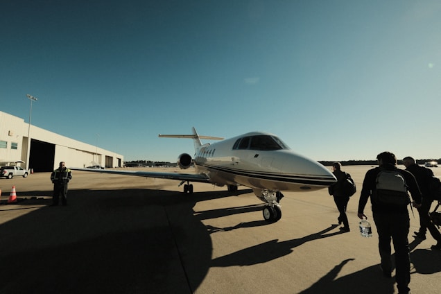 Several people walk towards a sleek private jet parked on an open tarmac. The sky is clear and blue, and the sun casts long shadows across the ground. A large hangar and some vehicles are visible in the background, indicating an airport or airfield setting.
