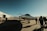 Several people walk towards a sleek private jet parked on an open tarmac. The sky is clear and blue, and the sun casts long shadows across the ground. A large hangar and some vehicles are visible in the background, indicating an airport or airfield setting.