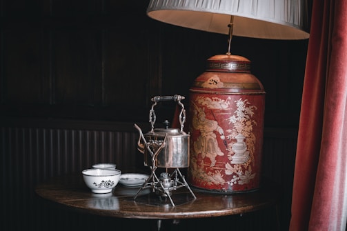 A vintage aesthetic featuring an ornate red tea canister with intricate gold designs, a decorative silver kettle, and two white bowls with black patterns, all set on a polished wooden table. The scene is accented by a gold-toned table lamp with a white shade and a heavy curtain in a dimly lit room.
