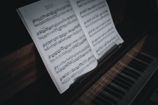 A close-up view of a piano keyboard with an open sheet of classical music. The sheet music rests on top of the piano, showing notes and musical notations in detail. The lighting creates a slightly moody and intimate atmosphere, with shadows enhancing the texture of the wood and keys.