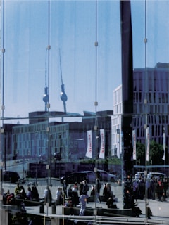 A glass facade reflects an urban scene, including a skyline with a tall tower featuring a spherical top. The reflection reveals several modern buildings and a street bustling with people walking and gathering. Vehicles are also visible on the road, and there are banners with text.