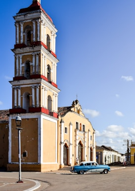 A historic church with a tall bell tower featuring red and white accents. The building has a weathered look, indicative of its age. There is a vintage blue car parked in front of the church. The skies are clear and blue, suggesting daytime with sunny weather.