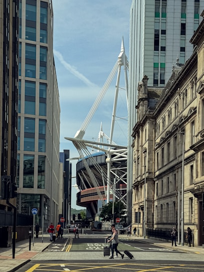 A city street flanked by modern and historic buildings with a large stadium featuring prominent white structural elements in the background. A few pedestrians, including one pulling a suitcase, walk along the road and sidewalk.