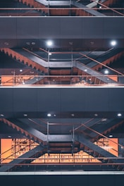 Multiple levels of open floors in a modern library are visible, with metal staircases and glass railings. Bookshelves filled with books are illuminated by warm lighting, and there are geometrical patterns created by the architectural structure.