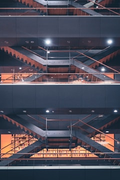 Multiple levels of open floors in a modern library are visible, with metal staircases and glass railings. Bookshelves filled with books are illuminated by warm lighting, and there are geometrical patterns created by the architectural structure.