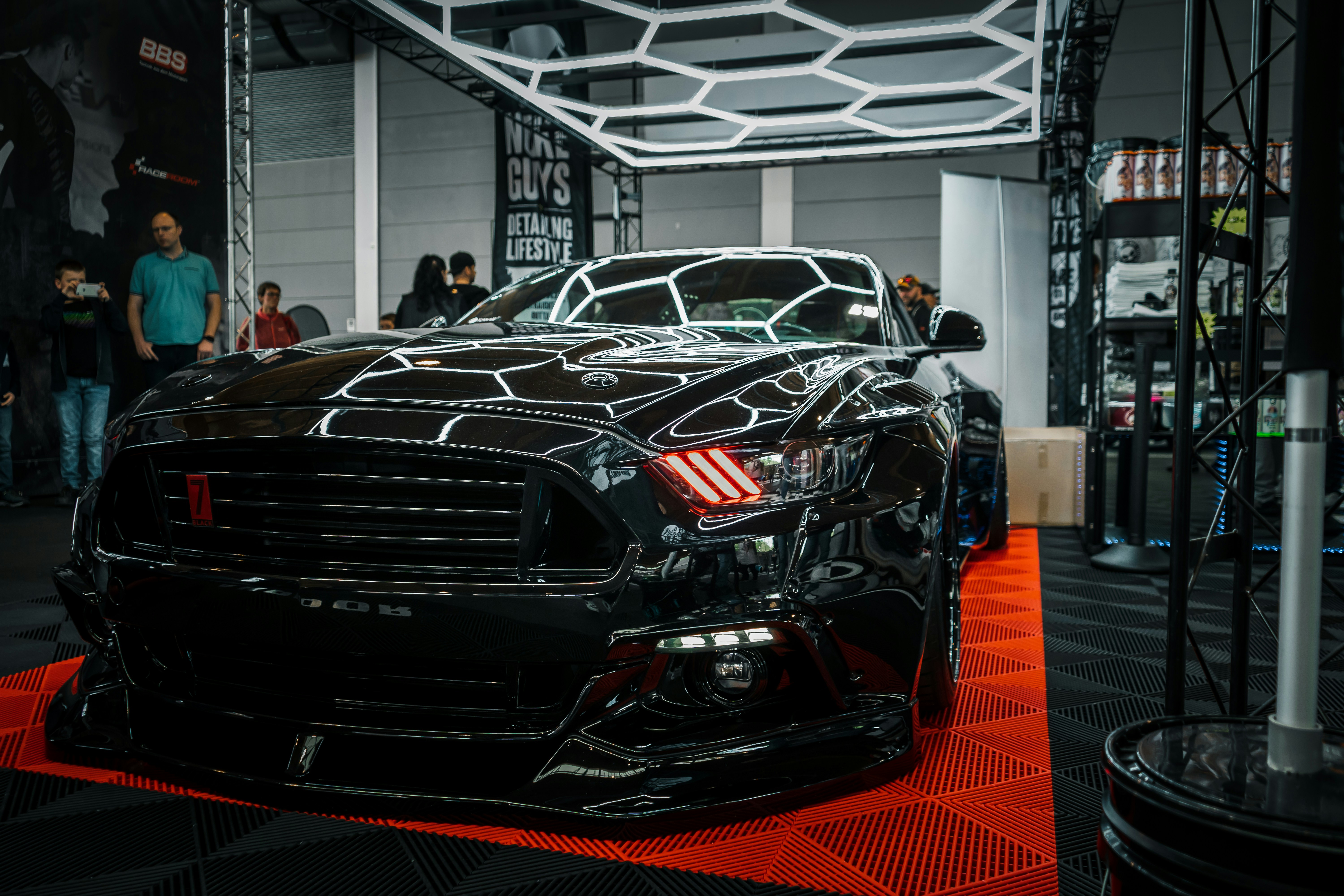 A sleek black sports car is prominently displayed indoors, surrounded by a few onlookers and car enthusiasts. The car's shiny exterior reflects the surrounding geometric lighting structures. A sign in the background suggests it's part of an automotive detailing event.