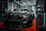 A sleek black sports car is prominently displayed indoors, surrounded by a few onlookers and car enthusiasts. The car's shiny exterior reflects the surrounding geometric lighting structures. A sign in the background suggests it's part of an automotive detailing event.