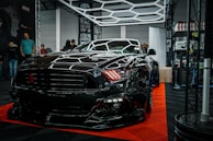 A sleek black sports car is prominently displayed indoors, surrounded by a few onlookers and car enthusiasts. The car's shiny exterior reflects the surrounding geometric lighting structures. A sign in the background suggests it's part of an automotive detailing event.