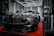 A sleek black sports car is prominently displayed indoors, surrounded by a few onlookers and car enthusiasts. The car's shiny exterior reflects the surrounding geometric lighting structures. A sign in the background suggests it's part of an automotive detailing event.