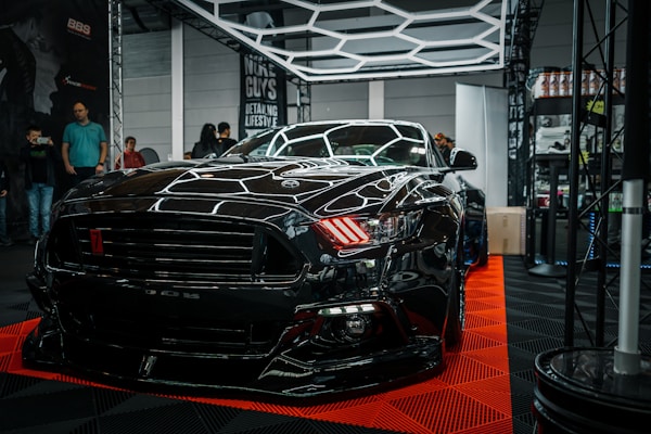 A sleek black sports car is prominently displayed indoors, surrounded by a few onlookers and car enthusiasts. The car's shiny exterior reflects the surrounding geometric lighting structures. A sign in the background suggests it's part of an automotive detailing event.