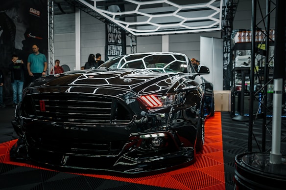 A sleek black sports car is prominently displayed indoors, surrounded by a few onlookers and car enthusiasts. The car's shiny exterior reflects the surrounding geometric lighting structures. A sign in the background suggests it's part of an automotive detailing event.