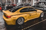 A yellow sports car with Deutsche Post branding and various sponsor logos such as Shell and MAHLE is displayed indoors with multiple other cars and people in the background. The car has large, shiny alloy wheels and is parked on a glossy, dark floor in a showroom or exhibition hall.