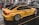 A yellow sports car with Deutsche Post branding and various sponsor logos such as Shell and MAHLE is displayed indoors with multiple other cars and people in the background. The car has large, shiny alloy wheels and is parked on a glossy, dark floor in a showroom or exhibition hall.