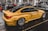 A yellow sports car with Deutsche Post branding and various sponsor logos such as Shell and MAHLE is displayed indoors with multiple other cars and people in the background. The car has large, shiny alloy wheels and is parked on a glossy, dark floor in a showroom or exhibition hall.
