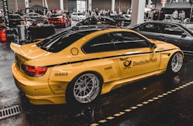 A yellow sports car with Deutsche Post branding and various sponsor logos such as Shell and MAHLE is displayed indoors with multiple other cars and people in the background. The car has large, shiny alloy wheels and is parked on a glossy, dark floor in a showroom or exhibition hall.