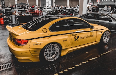 A yellow sports car with Deutsche Post branding and various sponsor logos such as Shell and MAHLE is displayed indoors with multiple other cars and people in the background. The car has large, shiny alloy wheels and is parked on a glossy, dark floor in a showroom or exhibition hall.