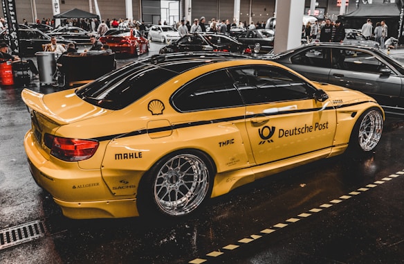 A yellow sports car with Deutsche Post branding and various sponsor logos such as Shell and MAHLE is displayed indoors with multiple other cars and people in the background. The car has large, shiny alloy wheels and is parked on a glossy, dark floor in a showroom or exhibition hall.