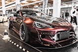 A modified sports car with a glossy, dark metallic finish is parked indoors at an automotive event. The car is equipped with a wide body kit, large wheels, and custom front modifications. Several other cars and people are visible in the background, with banners and lights suggesting a car show setting.