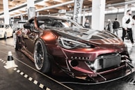 A modified sports car with a glossy, dark metallic finish is parked indoors at an automotive event. The car is equipped with a wide body kit, large wheels, and custom front modifications. Several other cars and people are visible in the background, with banners and lights suggesting a car show setting.