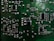 A close-up view of a green printed circuit board (PCB) with multiple electronic components such as resistors, capacitors, and integrated circuits. The PCB features intricate pathways and metallic connections, showcasing a detailed arrangement of electronic circuitry.