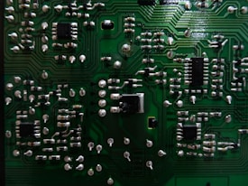 A close-up view of a green printed circuit board (PCB) with multiple electronic components such as resistors, capacitors, and integrated circuits. The PCB features intricate pathways and metallic connections, showcasing a detailed arrangement of electronic circuitry.