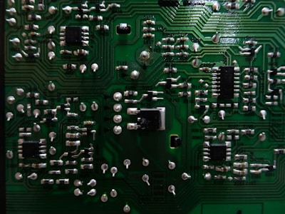 A close-up view of a green printed circuit board (PCB) with multiple electronic components such as resistors, capacitors, and integrated circuits. The PCB features intricate pathways and metallic connections, showcasing a detailed arrangement of electronic circuitry.