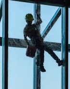A person is suspended outside a tall building, cleaning a large window. The silhouette of the person, wearing a safety harness and a hard hat, is clearly visible against the blue sky. The person holds a bucket in one hand while cleaning the glass surface.