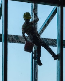 A person is suspended outside a tall building, cleaning a large window. The silhouette of the person, wearing a safety harness and a hard hat, is clearly visible against the blue sky. The person holds a bucket in one hand while cleaning the glass surface.