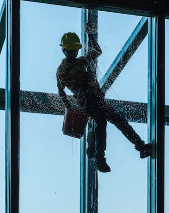 A person is suspended outside a tall building, cleaning a large window. The silhouette of the person, wearing a safety harness and a hard hat, is clearly visible against the blue sky. The person holds a bucket in one hand while cleaning the glass surface.