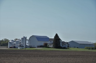 A rural landscape featuring a farmhouse surrounded by silos and large barns. The image presents a clear, open sky, green lawns, and trees lining the background, with freshly tilled soil in the foreground.