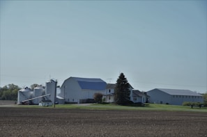 A rural landscape featuring a farmhouse surrounded by silos and large barns. The image presents a clear, open sky, green lawns, and trees lining the background, with freshly tilled soil in the foreground.