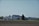 A rural landscape featuring a farmhouse surrounded by silos and large barns. The image presents a clear, open sky, green lawns, and trees lining the background, with freshly tilled soil in the foreground.