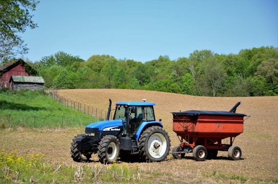 A blue tractor is positioned in a freshly plowed field, pulling a red trailer. A rustic red barn sits in the background surrounded by lush green trees, and the sky is a clear, bright blue.