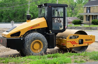 A yellow SANY SSR120C-8 road roller is parked on a dirt road. The background consists of a lush green landscape with trees and a yellow house. The roller has a large cylindrical drum used for compacting surfaces.