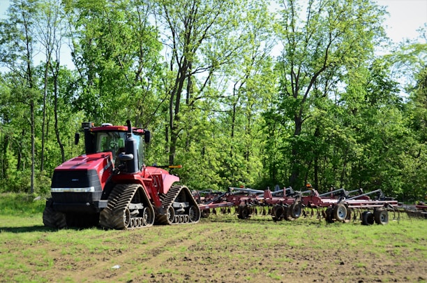 A large red agricultural tractor with caterpillar tracks is parked on a field, surrounded by tall green trees. It is attached to a plow or farming equipment, ready for work. The setting is rural and peaceful.
