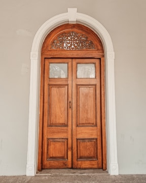 A wooden door with an ornate arch design at the top, framed by white decorative molding against a light gray wall. The door has two panels and features a small keyhole.