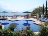 A serene outdoor scene featuring a luxurious pool with clear blue water surrounded by vibrant plants and flowers. There are several lounge chairs and tables with umbrellas on a stone patio overlooking a large body of water, with mountainous terrain visible in the distance. The area is landscaped with potted plants and decorative elements, creating a peaceful, vacation-like atmosphere.