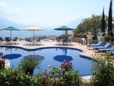 A serene outdoor scene featuring a luxurious pool with clear blue water surrounded by vibrant plants and flowers. There are several lounge chairs and tables with umbrellas on a stone patio overlooking a large body of water, with mountainous terrain visible in the distance. The area is landscaped with potted plants and decorative elements, creating a peaceful, vacation-like atmosphere.