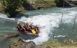 A group of five people wearing helmets and life jackets are paddling in a red inflatable raft through a fast-moving river with turbulent white water. The surrounding area features green foliage and rocks. A pair of poles are placed on either side of the waterway, indicating a slalom course.