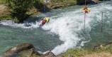 A person in a helmet and life jacket maneuvers a kayak through fast-moving rapids, navigating between poles in a river surrounded by greenery. The water is white and frothy, indicating the strength of the current.