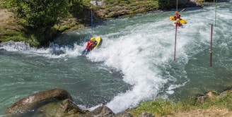 A person in a helmet and life jacket maneuvers a kayak through fast-moving rapids, navigating between poles in a river surrounded by greenery. The water is white and frothy, indicating the strength of the current.