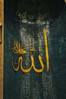 Large Arabic calligraphy in golden color is displayed on a background of teal mosaic tiles. The script is artistically ornate and dominates the visual plane.
