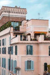 A multi-story building with an elegant architectural design featuring light pastel-colored walls and numerous windows with blue shutters. The roof is adorned with greenery, and a balcony with plants hints at a touch of nature amidst urban living. Ornate detailing is present around the windows, and the topmost portion has a more rustic brick appearance with decorative elements.
