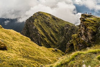 A stunning landscape featuring a rugged mountain with vibrant green grass and rocky terrain. The sky is filled with dramatic clouds, and a group of people stands on the edge of a cliff, creating a sense of scale and adventure.