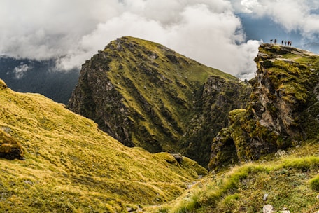 A stunning landscape featuring a rugged mountain with vibrant green grass and rocky terrain. The sky is filled with dramatic clouds, and a group of people stands on the edge of a cliff, creating a sense of scale and adventure.