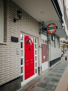 A street view showing a brick building with a bright red door. A sign above reads 'Shayegan' and there are modern fixtures such as lantern-style lights. The sidewalk is tiled with alternating patterns and colors.