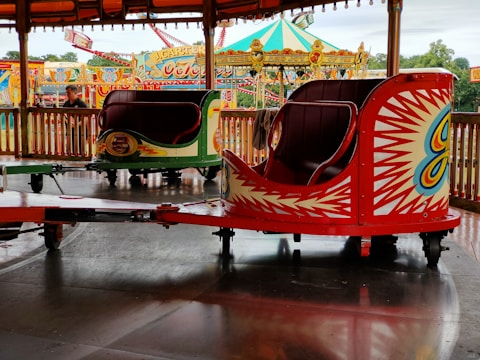 A vibrant amusement park scene featuring colorful spinning ride cars with bold geometric patterns. The foreground showcases a red ride car with white and yellow starburst designs, while the background includes a carousel tent and additional ride structures adorned with vivid colors and intricate lettering.