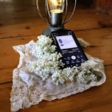 A smartphone rests on a wooden surface and displays a podcast episode, surrounded by a lace cloth and white lilac flowers. A decorative lantern with a glowing light bulb stands nearby.