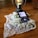 A smartphone rests on a wooden surface and displays a podcast episode, surrounded by a lace cloth and white lilac flowers. A decorative lantern with a glowing light bulb stands nearby.