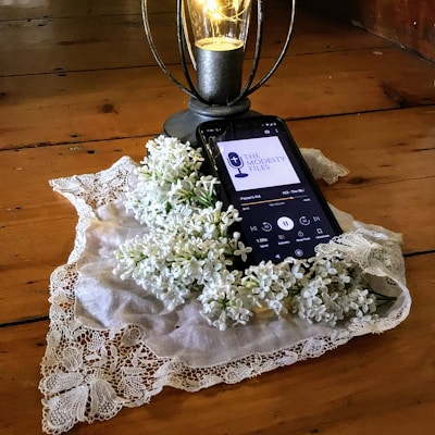 A smartphone rests on a wooden surface and displays a podcast episode, surrounded by a lace cloth and white lilac flowers. A decorative lantern with a glowing light bulb stands nearby.